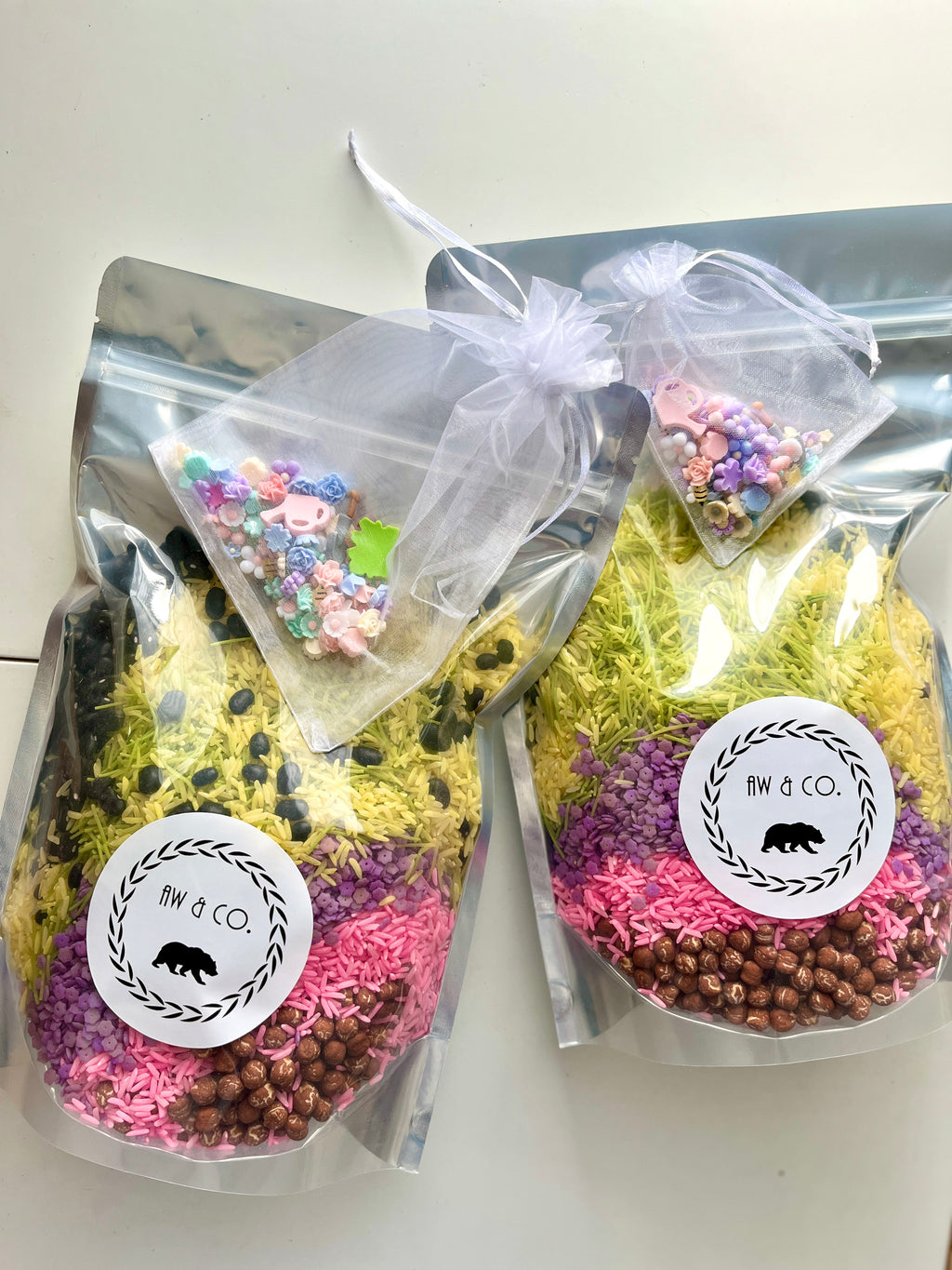 Two resealable bags filled with layered colored rice, small brown beads, and green pasta, each with a sheer pouch of pastel craft beads attached.