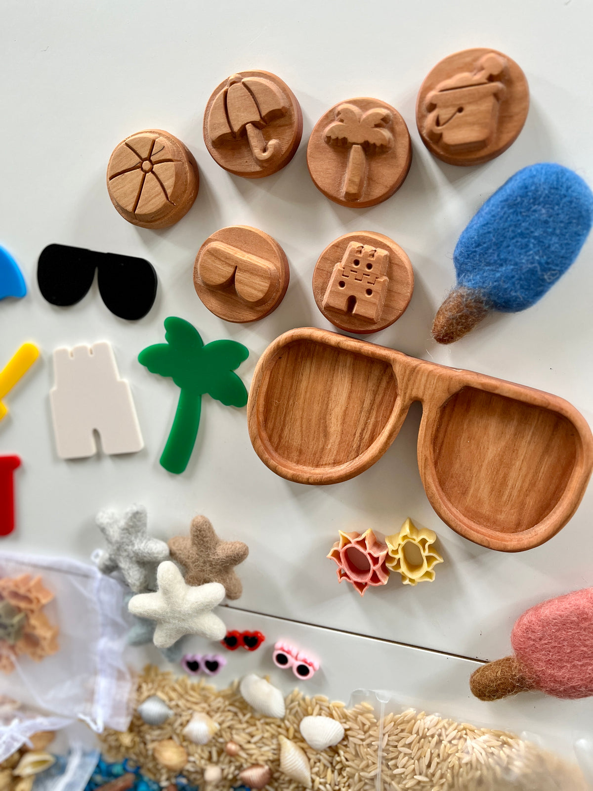 Wooden tokens with beach-themed carvings, felt shapes, plastic palm tree and sunglasses, and sensory materials on white surface