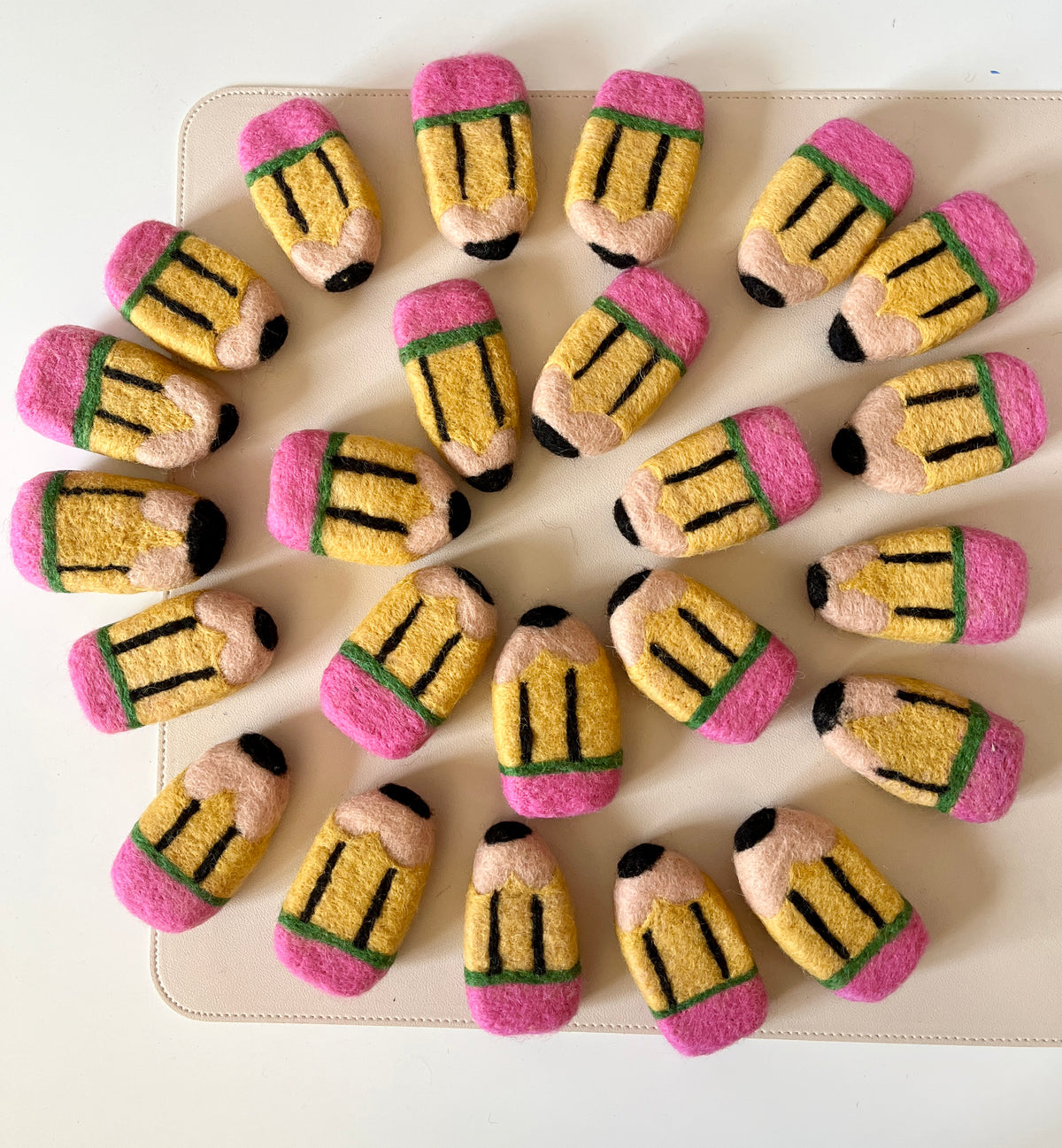 Twenty plush pencil-shaped toys with pink erasers and yellow bodies arranged in a circular pattern on a beige surface