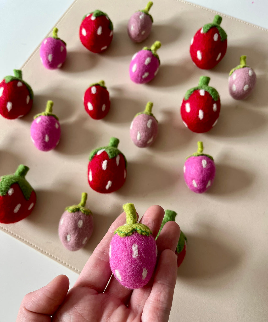 Hand holding a small purple and pink felt strawberry with scattered red, pink, and purple felt strawberries on a light surface