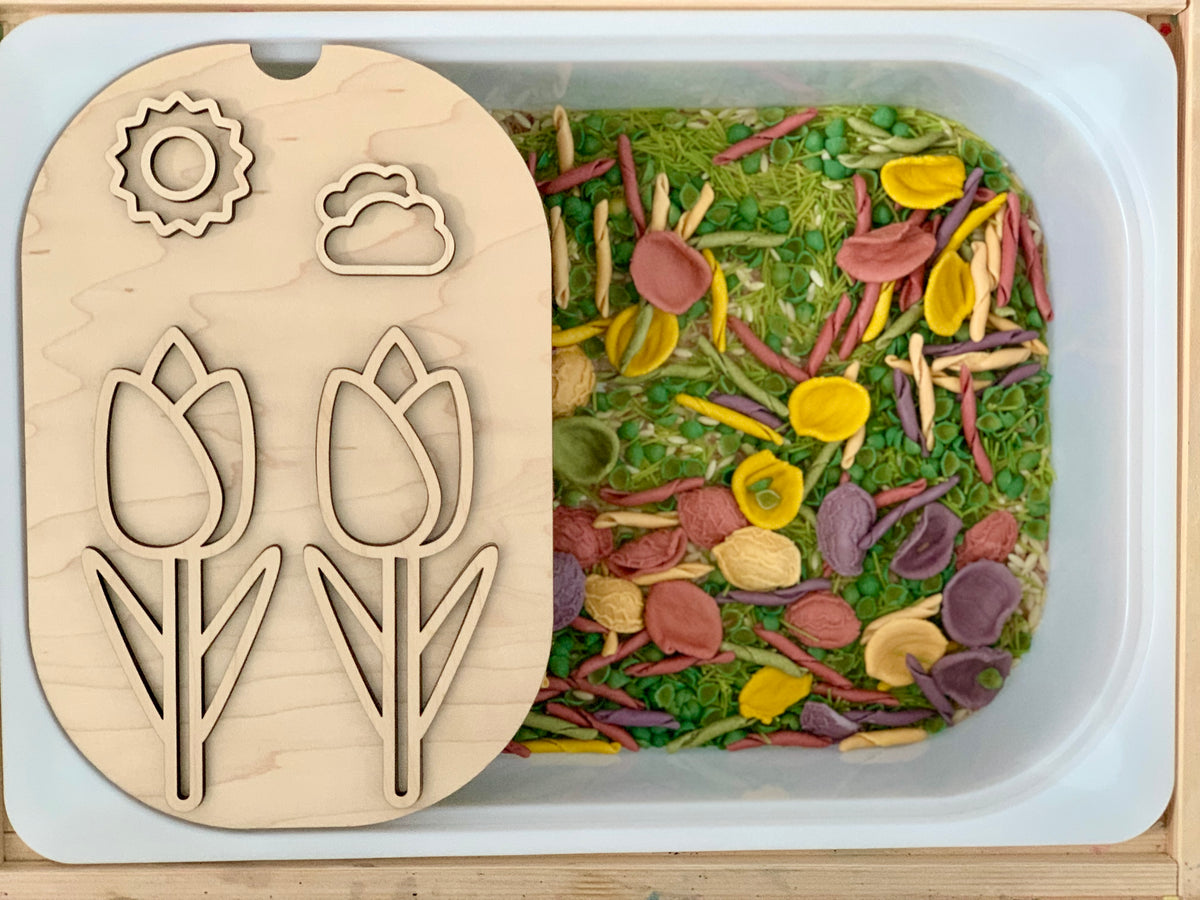 White sensory bin filled with green peas and colorful dried pasta with a wooden flower and weather shape puzzle lid on top