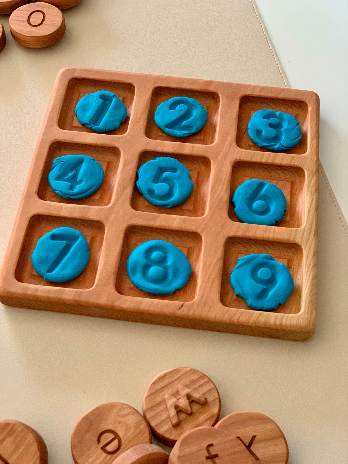 Wooden grid tray holding blue clay imprints of numbers 1 to 9 on a light surface with wooden letter tokens nearby