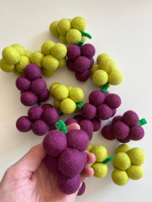 Hand holding a cluster of deep purple felt balls shaped like grapes among scattered yellow felt grape clusters on white background