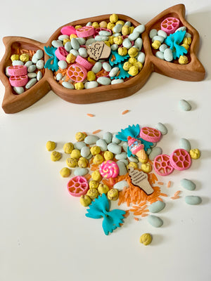 Wooden fish-shaped tray filled with colorful pasta, beans, and wooden ice cream and bow-shaped decorations, with some spilled pieces nearby