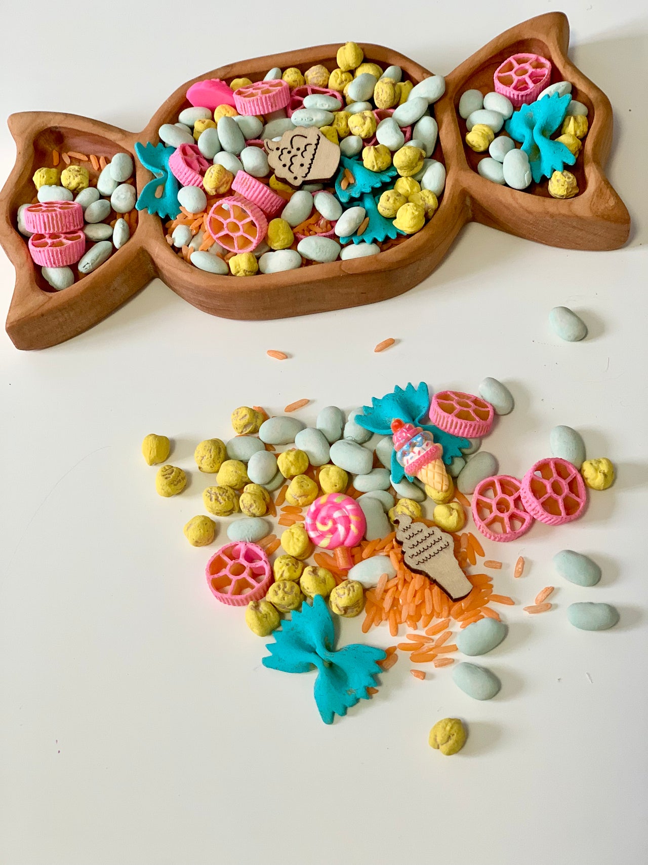 Wooden fish-shaped tray filled with colorful pasta, beans, and wooden ice cream and bow-shaped decorations, with some spilled pieces nearby