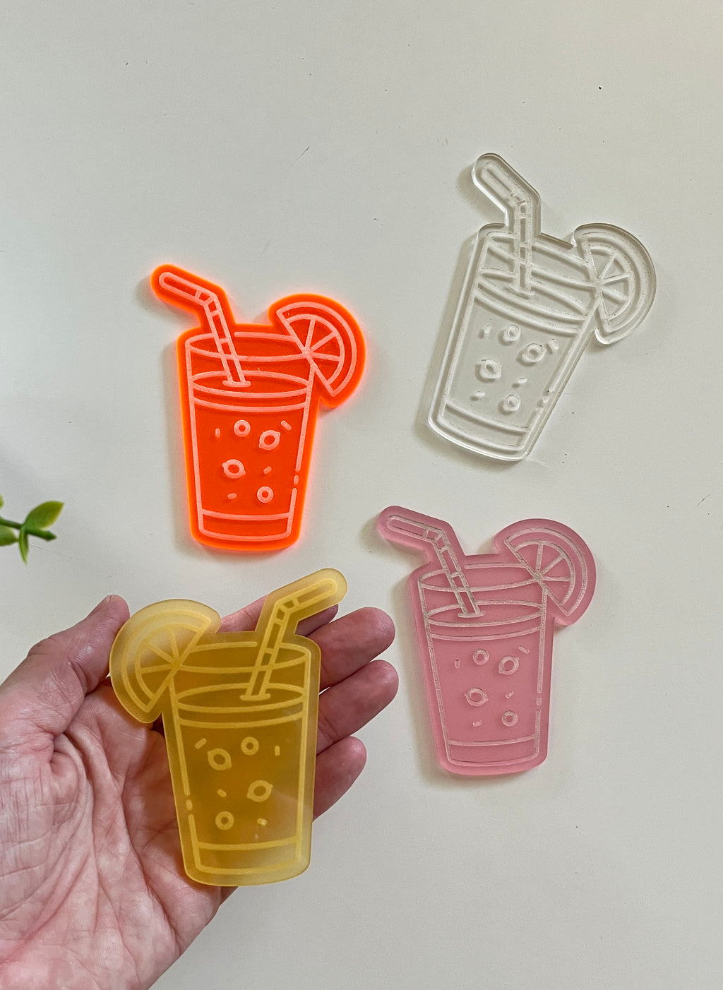 Hand holding a yellow translucent acrylic drink-shaped piece next to orange, pink, and clear similar pieces on white surface