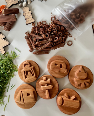 Wooden circular stamps with camping icons including lantern, camper, campfire, tent, signpost, and log beside brown play pieces and green grass strands