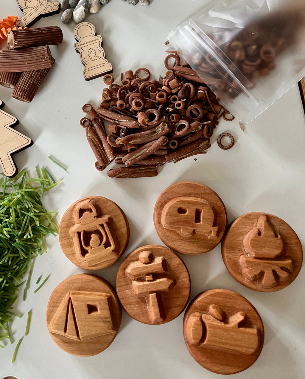 Wooden circular stamps with camping icons including lantern, camper, campfire, tent, signpost, and log beside brown play pieces and green grass strands