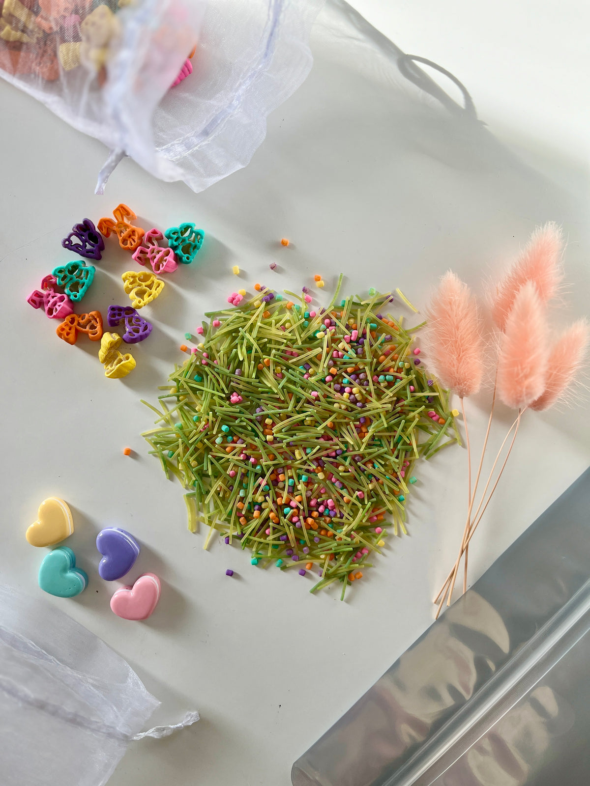 Colorful heart-shaped pasta pieces, green pasta strands with multicolored sprinkles, and pink fluffy dried flowers on white surface