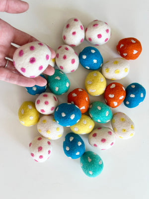 Hand holding a white felt egg with pink polka dots above scattered felt eggs in blue, yellow, orange, green, and white with colorful dots on white surface