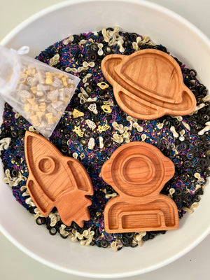 Wooden trays shaped like a rocket, astronaut helmet, and planet on black, white, and purple sensory rice mix with space-themed pasta pieces