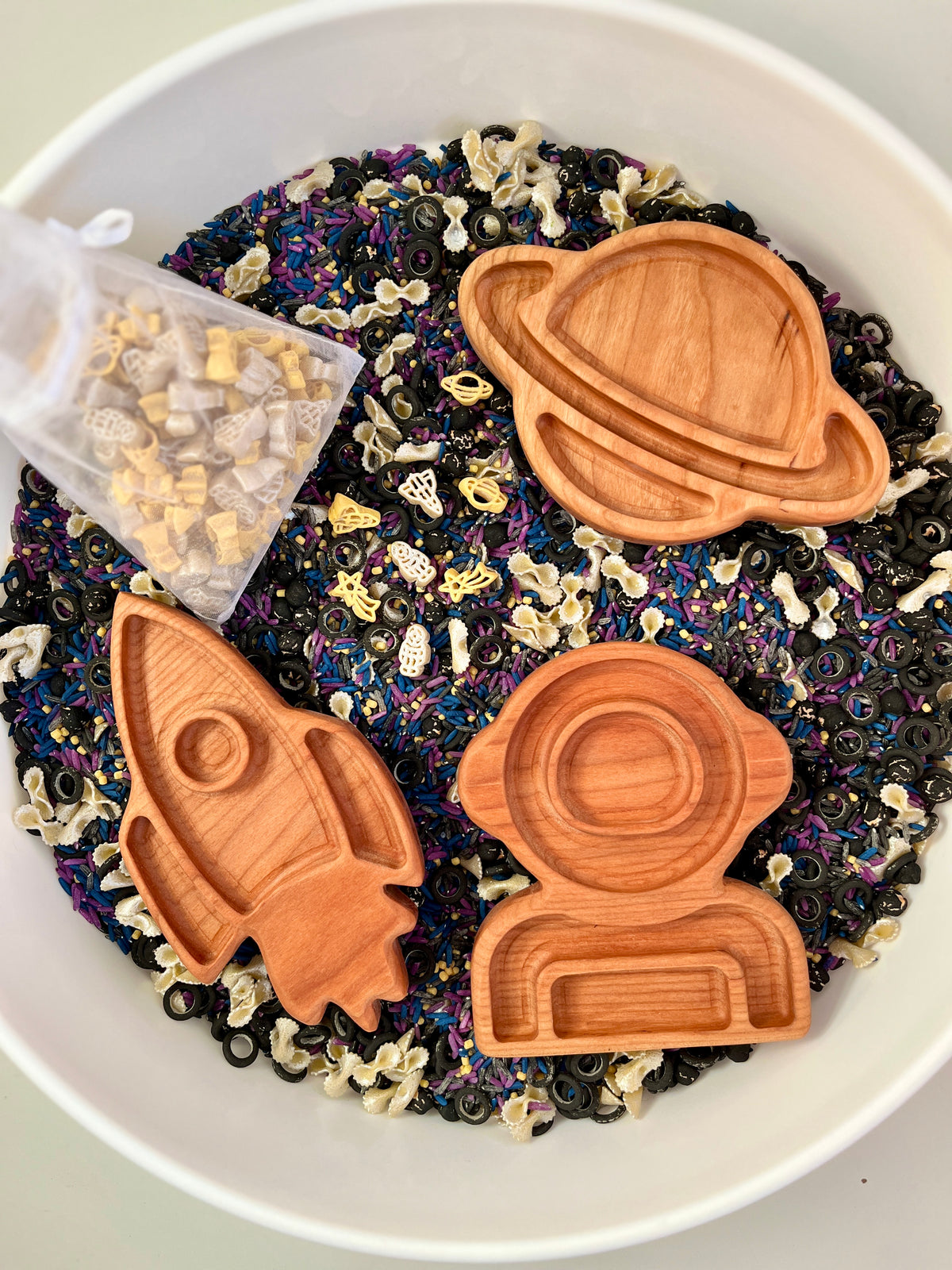 Wooden trays shaped like a rocket, astronaut helmet, and planet on black, white, and purple sensory rice mix with space-themed pasta pieces