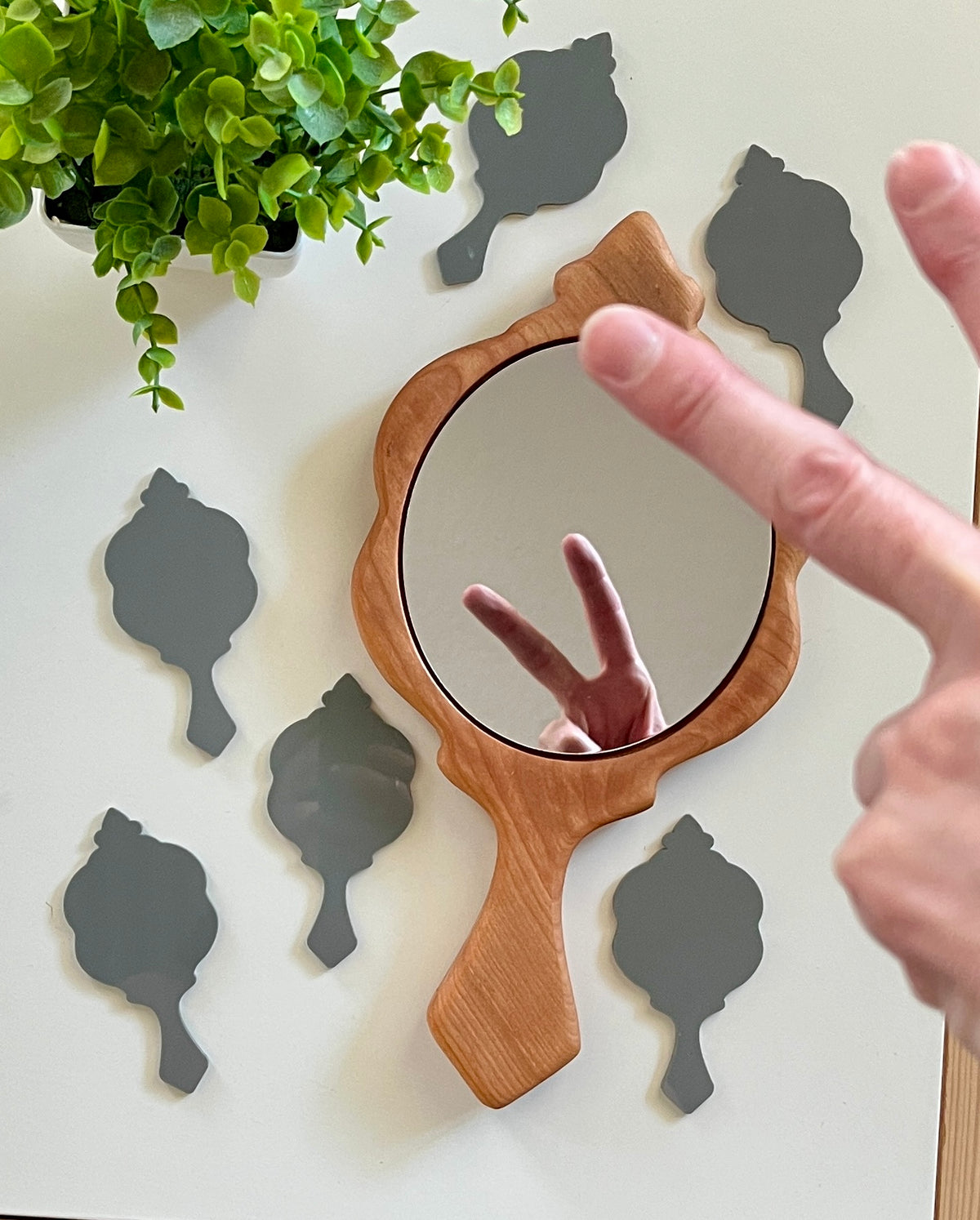 Wooden framed hand mirror reflecting fingers making a peace sign with gray mirror-shaped cutouts and a green plant nearby