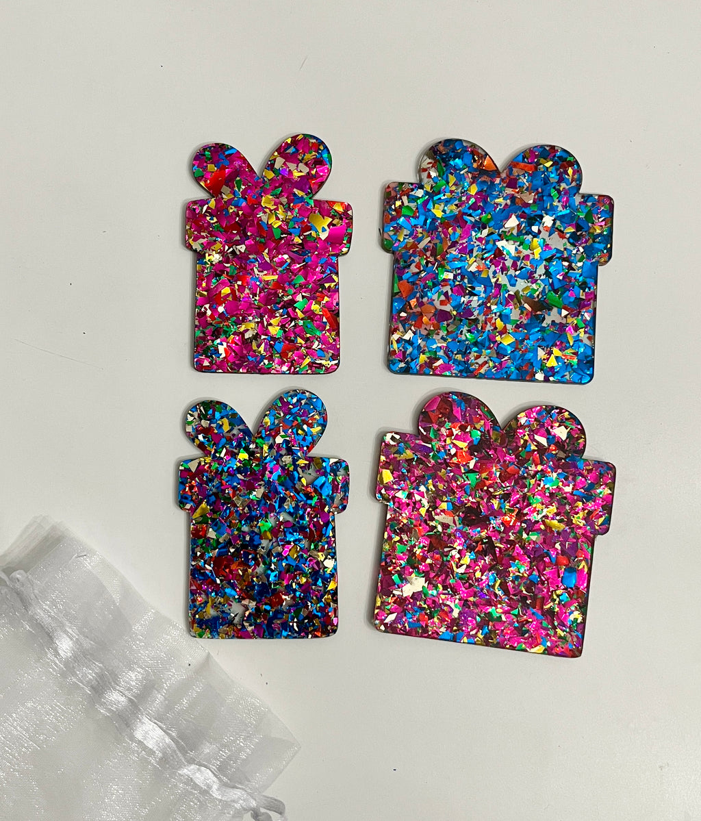 Four glittery present-shaped acrylic pieces in pink and blue with multicolored flakes on white background