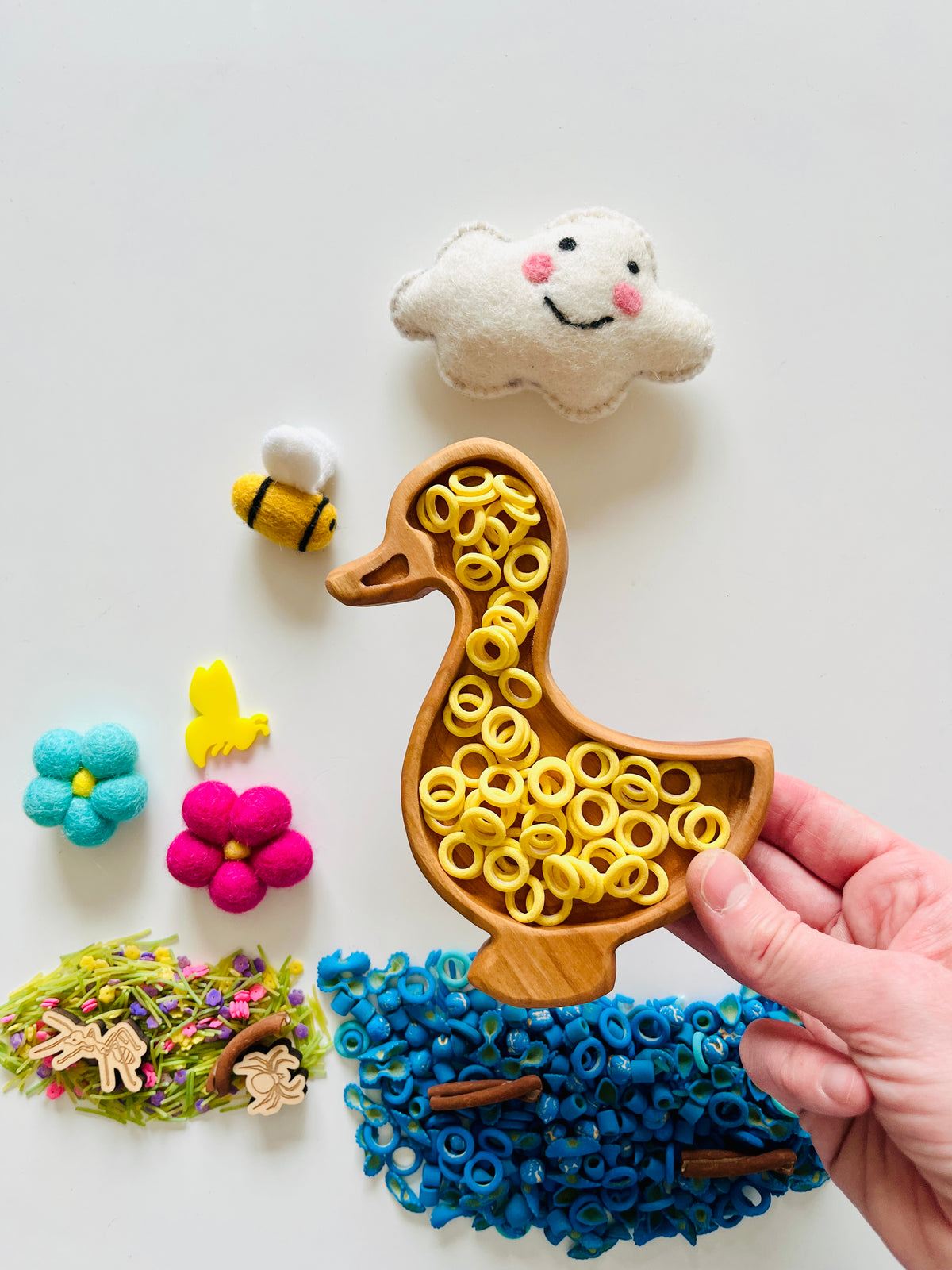 Hand holding wooden duck-shaped tray filled with yellow ring-shaped pasta beside felt flowers, bees, and a smiling cloud on white background