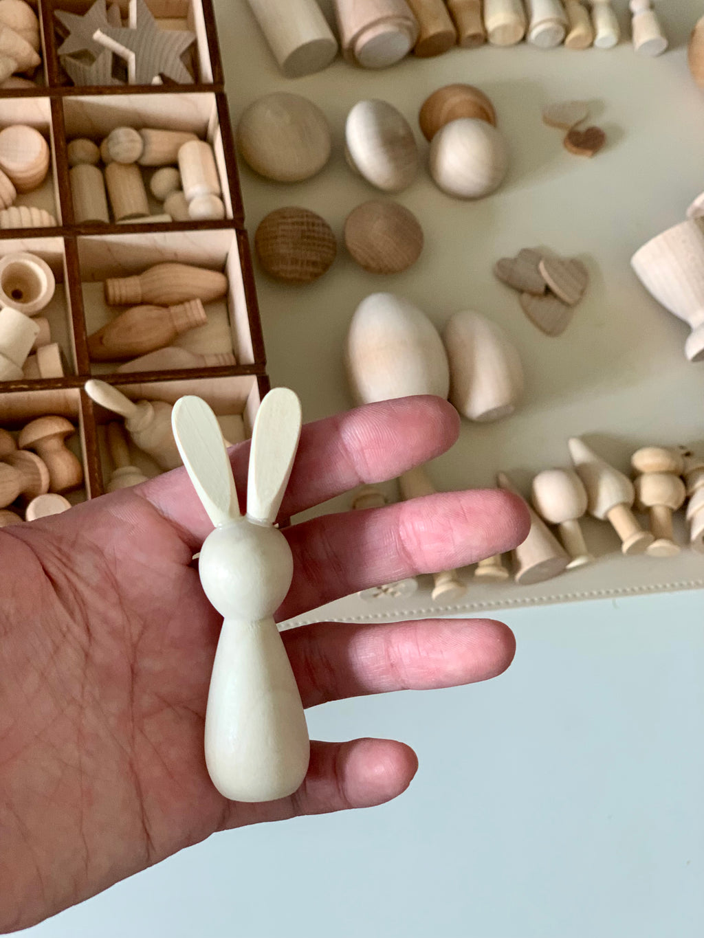 Hand holding smooth, unfinished wooden bunny figure with assorted natural wood shapes and peg dolls in background