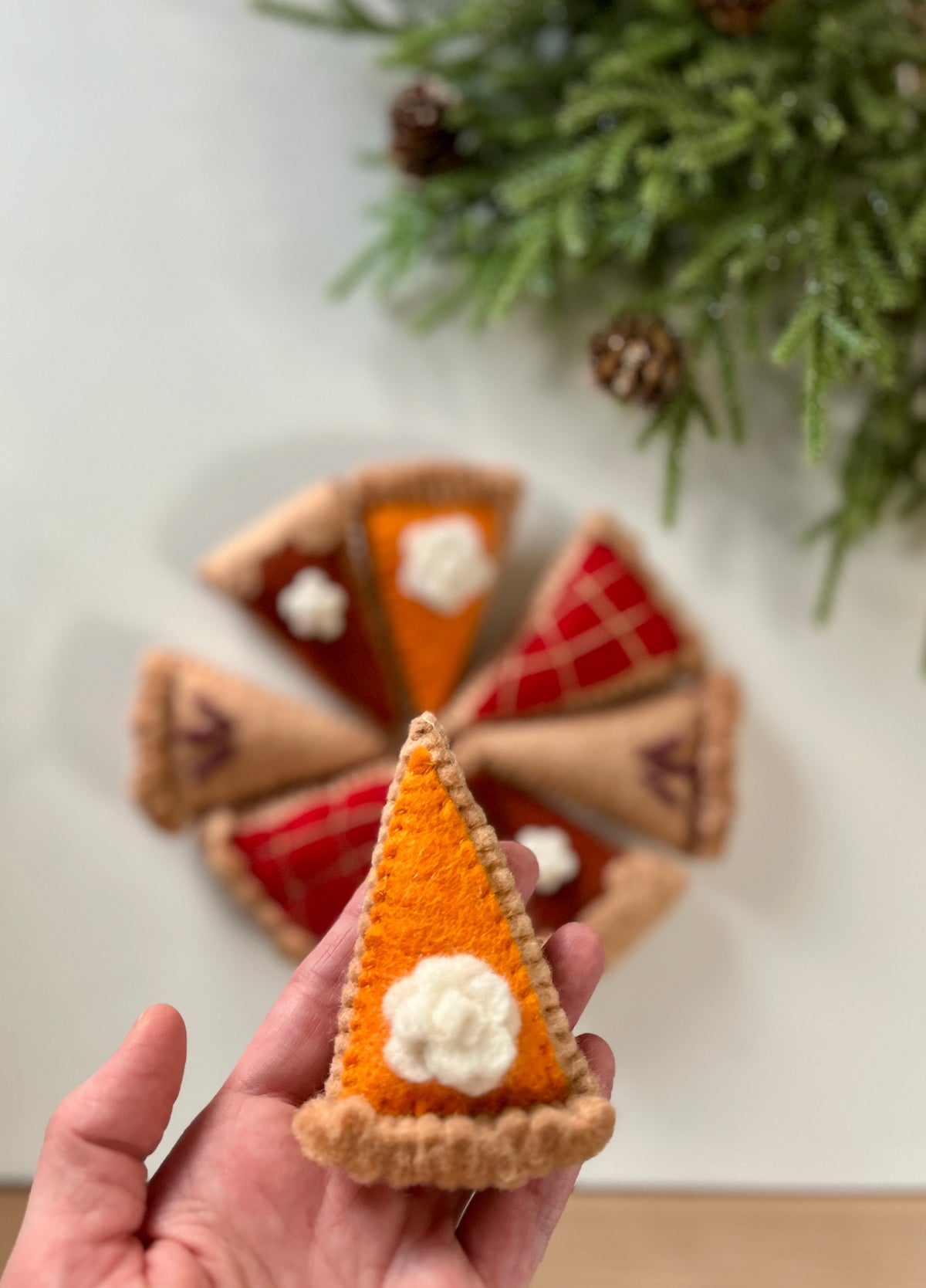 Felt pumpkin pie slice with whipped cream detail held in hand with other pie slice ornaments and greenery background