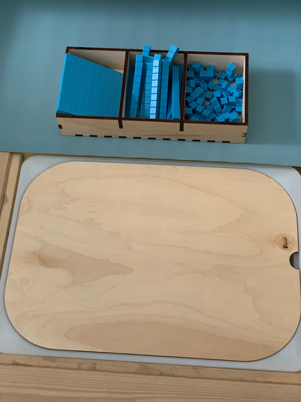 Wooden tray containing blue base ten blocks in hundreds, tens, and ones next to a blank wooden board on a blue surface