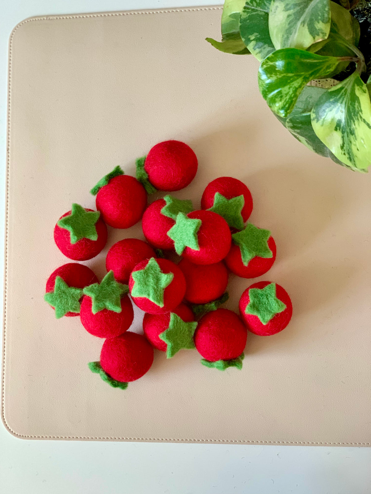 Pile of small red felt balls with green leaf tops arranged on a beige surface near a variegated green houseplant