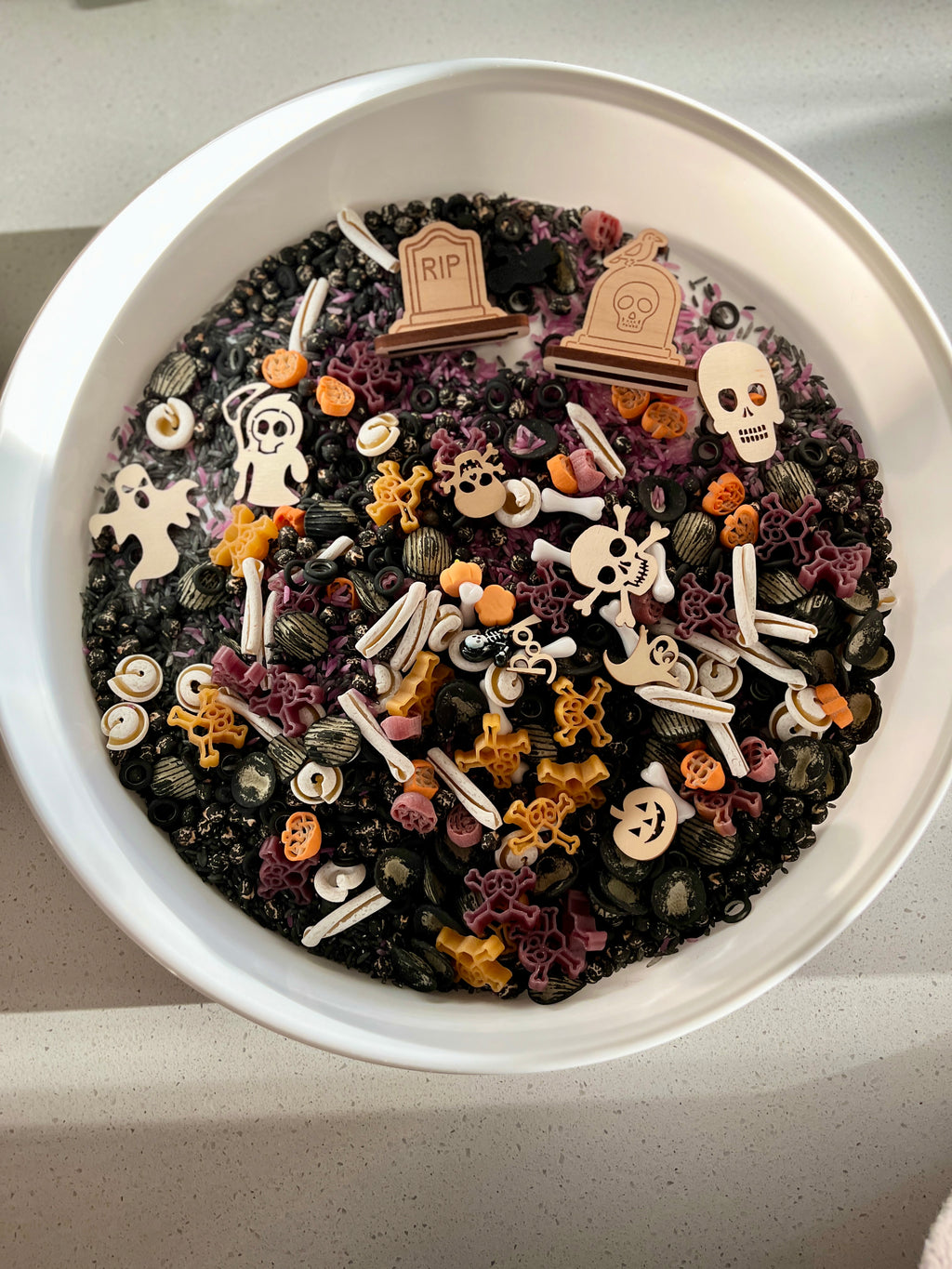 Round container filled with black, orange, purple, and white Halloween-themed pasta shapes including skulls and graves