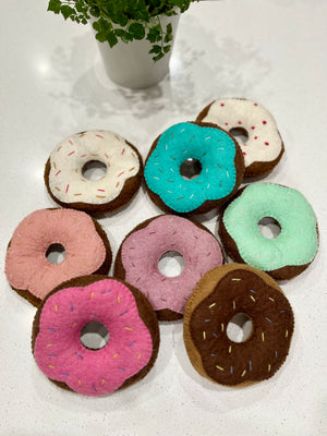 Seven handmade felt donuts in various pastel colors with stitched sprinkles arranged on a white surface near a potted plant