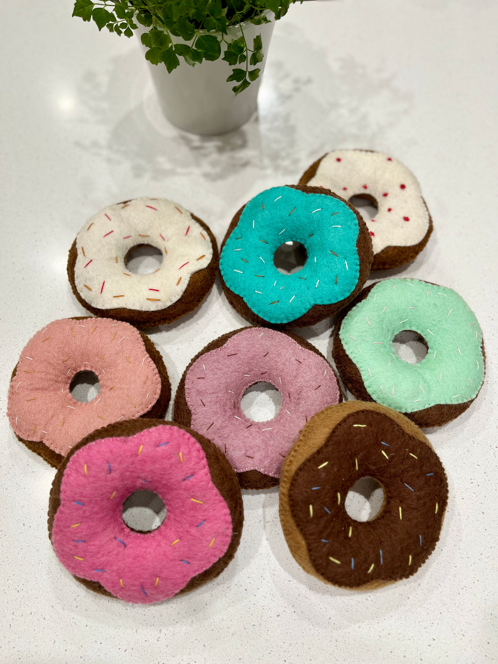 Seven handmade felt donuts in various pastel colors with stitched sprinkles arranged on a white surface near a potted plant