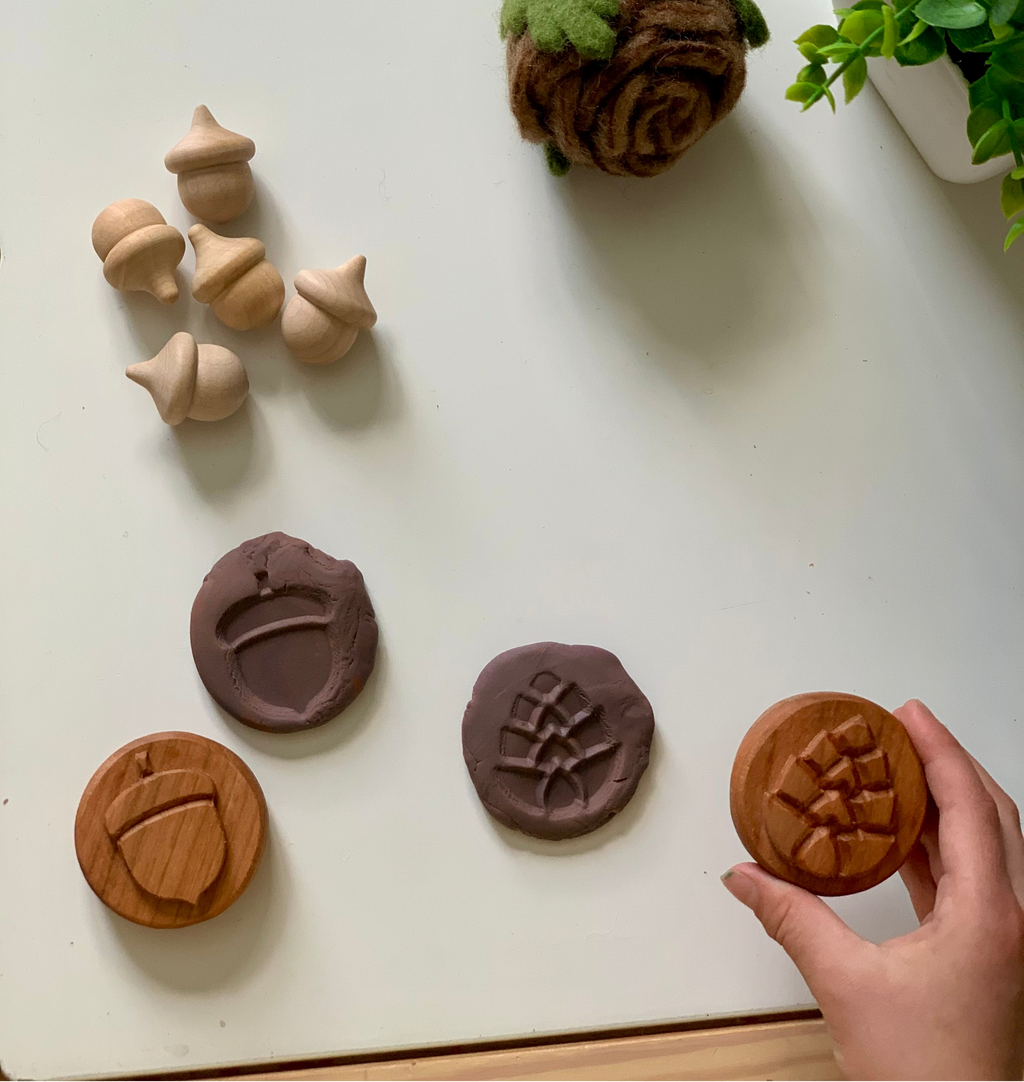 Hand holding round wooden stamp with pinecone design, two matching clay impressions, small wooden acorn figurines on white surface