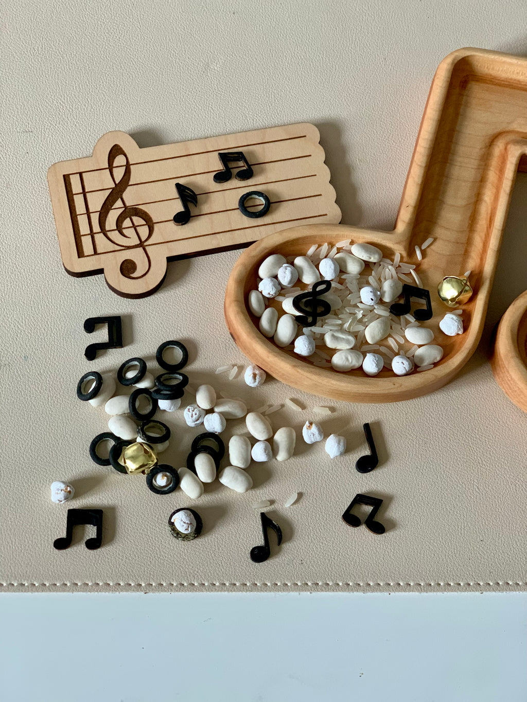 Wooden music note tray filled with white beans, rice, and black plastic musical note tokens on beige surface