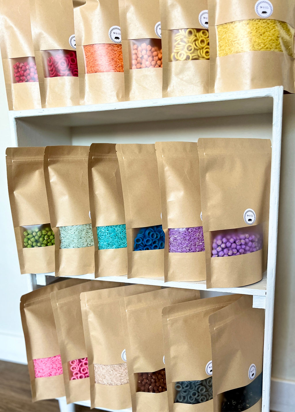 Assorted coloured sensory filler packs with rice, chickpeas, and pasta rings in kraft paper bags on white shelf