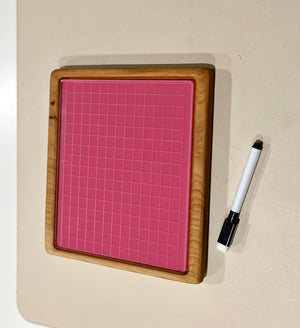 Wood-framed pink grid board with small squares and black and white dry erase marker on beige surface