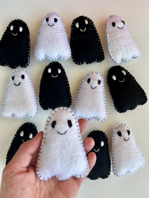 Hand holding a small white felt ghost with black stitched eyes and mouth among similar black and white felt ghosts on a light surface