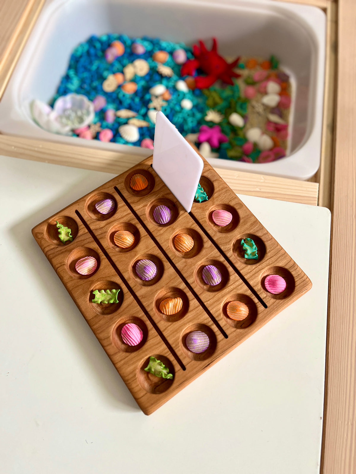 Wooden game board with rows of colorful seashell-shaped pieces and small green sea creature figures on a white table