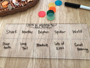 Clear board with handwritten animal names and adaptations, colorful counters, and dry erase marker on wooden surface