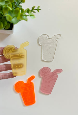 Four translucent acrylic shapes resembling drinks with straws and lemon slices in yellow, clear, orange, and pink colors on a white surface