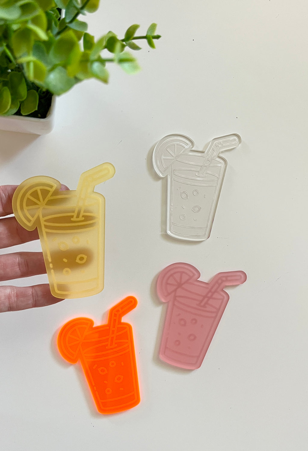 Four translucent acrylic shapes resembling drinks with straws and lemon slices in yellow, clear, orange, and pink colors on a white surface