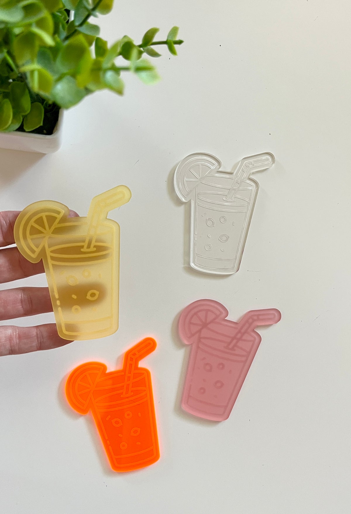 Four translucent acrylic shapes resembling drinks with straws and lemon slices in yellow, clear, orange, and pink colors on a white surface