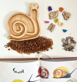 Wooden snail-shaped tray next to natural brown sensory materials, small snail toys, pebbles, and an open book about snails
