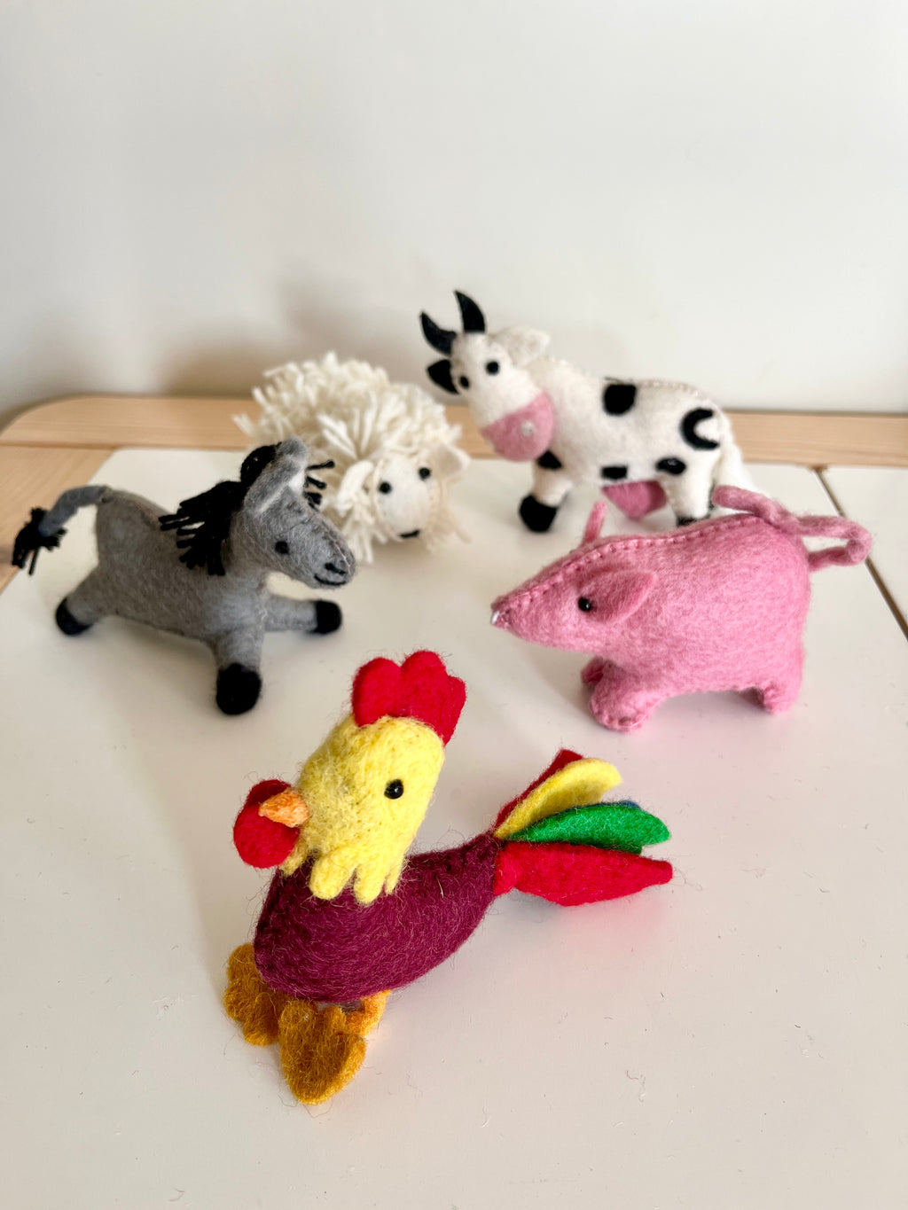 Six small handmade felt farm animal dolls including a rooster, pig, cow, donkey, and sheep on a light surface