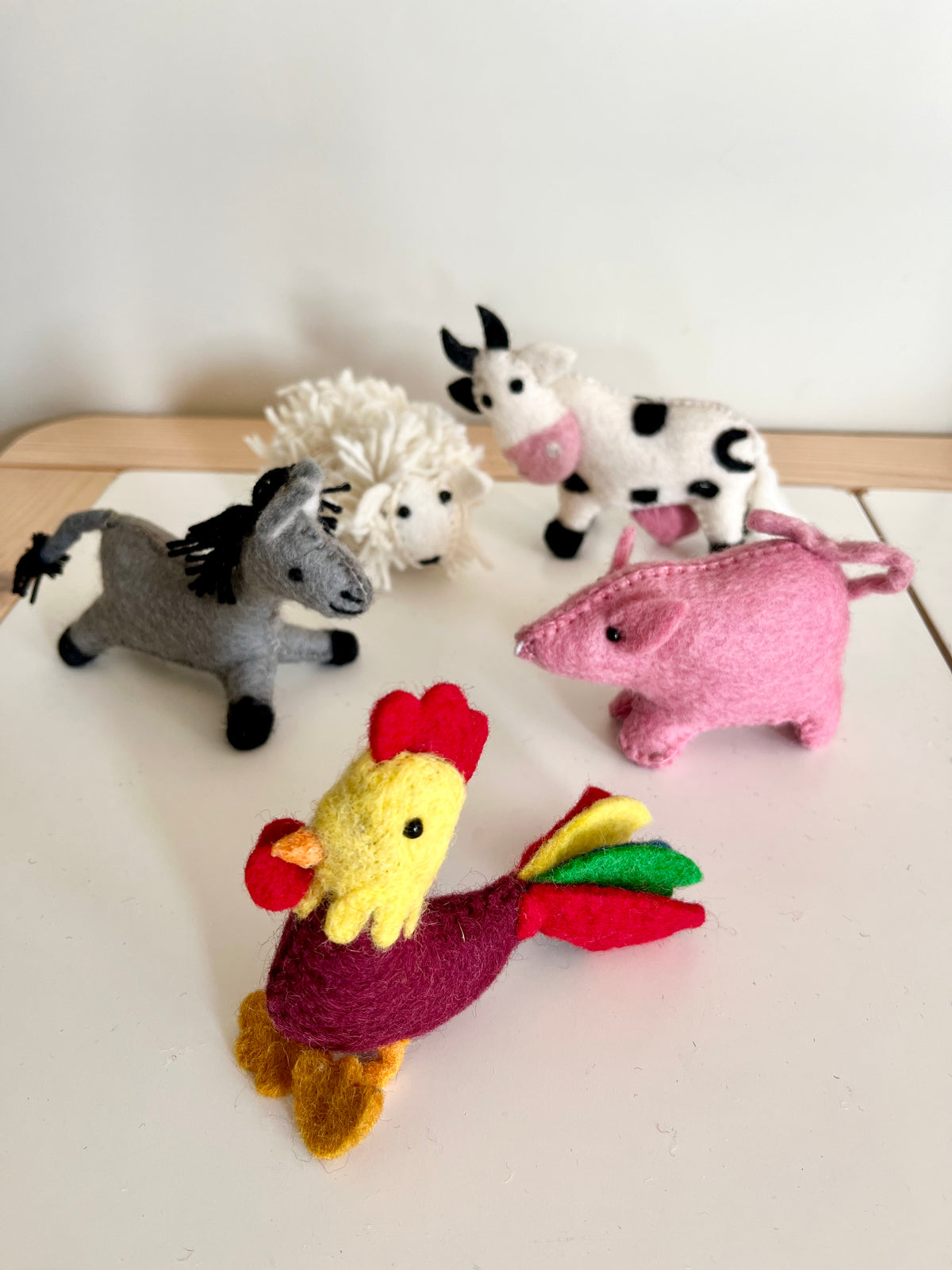 Six small handmade felt farm animal dolls including a rooster, pig, cow, donkey, and sheep on a light surface