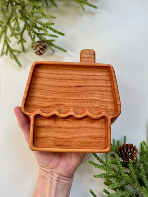 Hand holding a wooden tray shaped like a house with chimney, surrounded by pine branches and pine cones on white surface