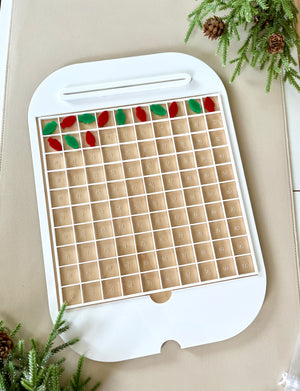 White frame with numbered wooden grid compartments and red and green leaf-shaped tokens on beige surface with pine branches