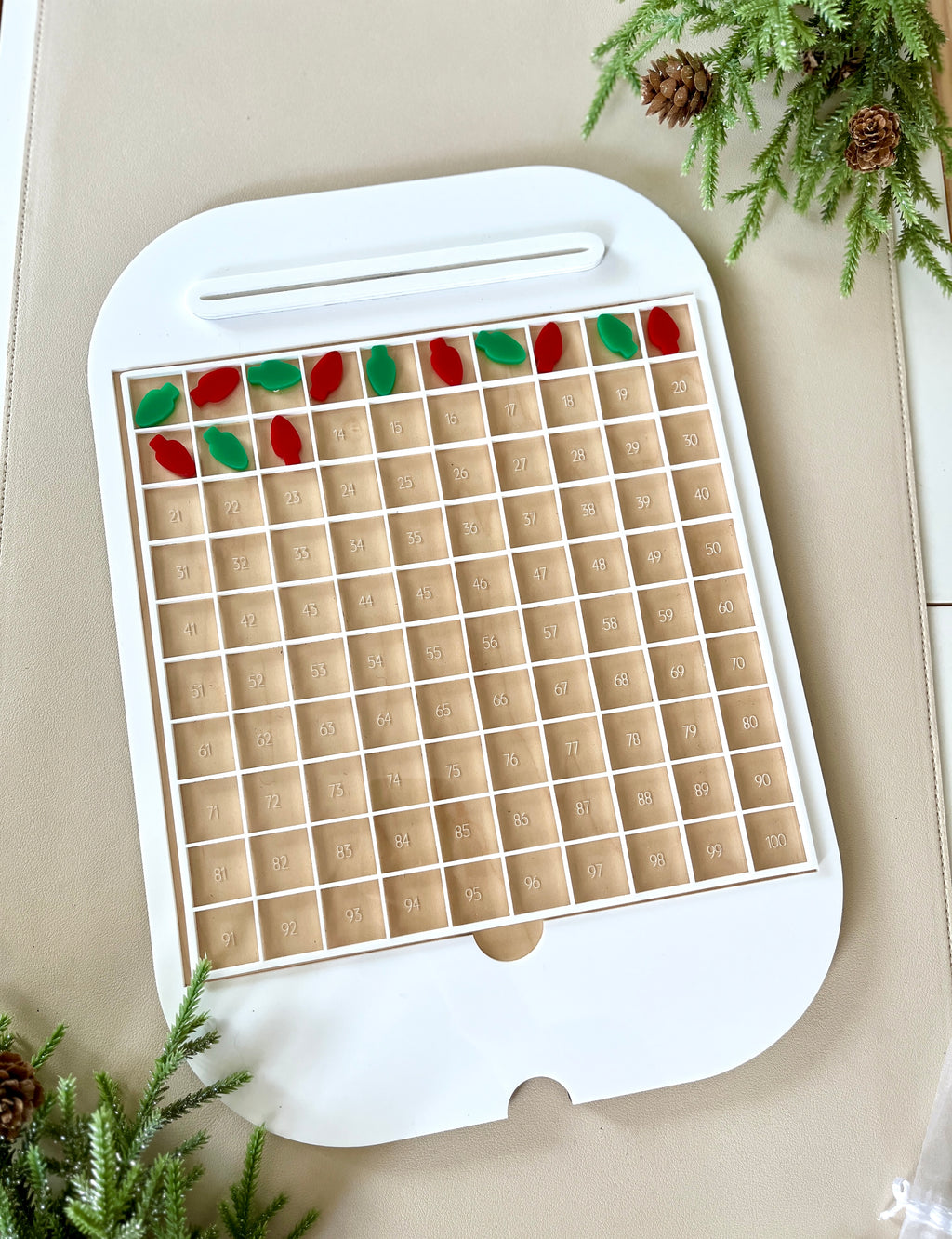 White frame with numbered wooden grid compartments and red and green leaf-shaped tokens on beige surface with pine branches
