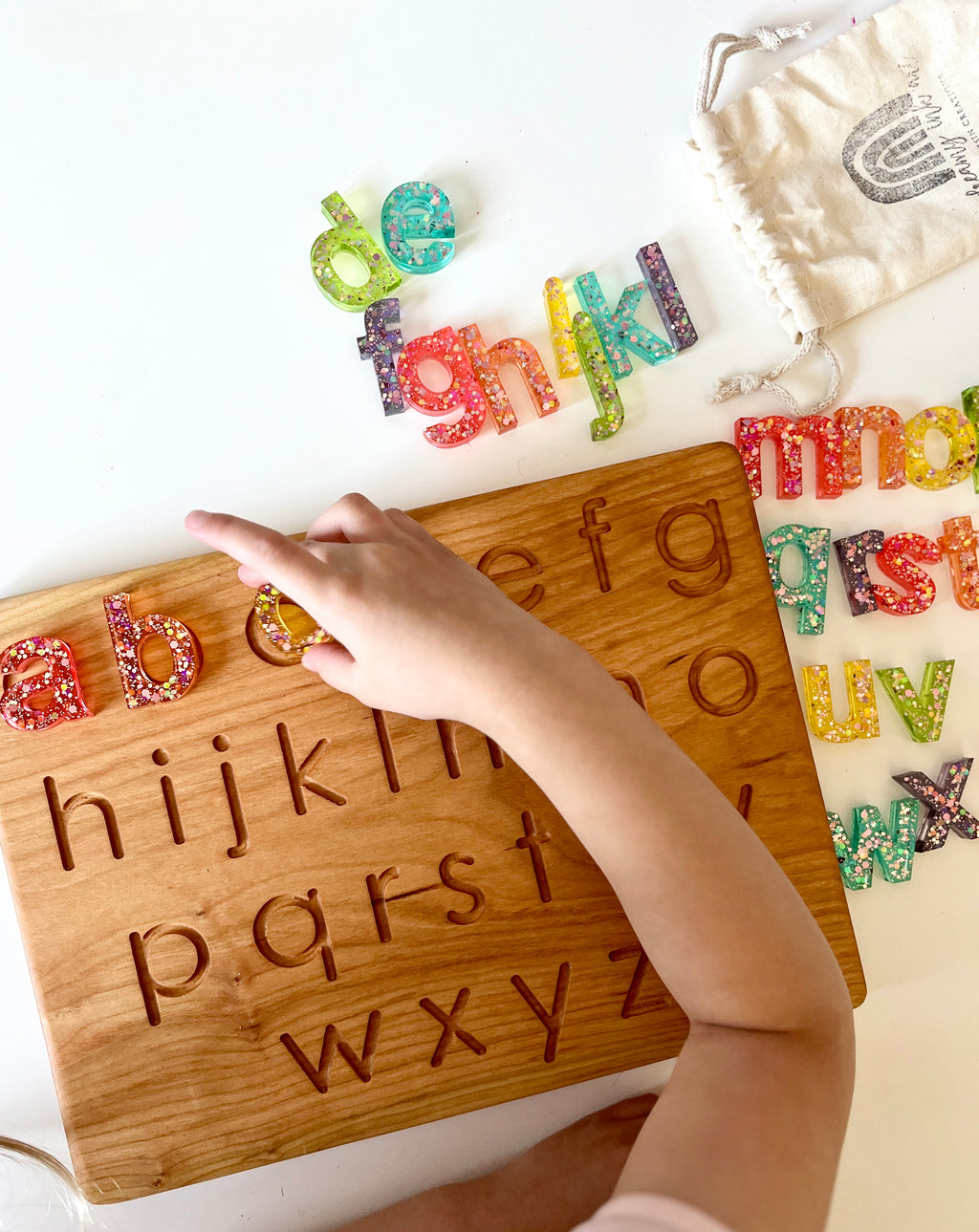 Child’s hand placing colorful resin lowercase letters onto matching carved wooden alphabet board with storage pouch nearby