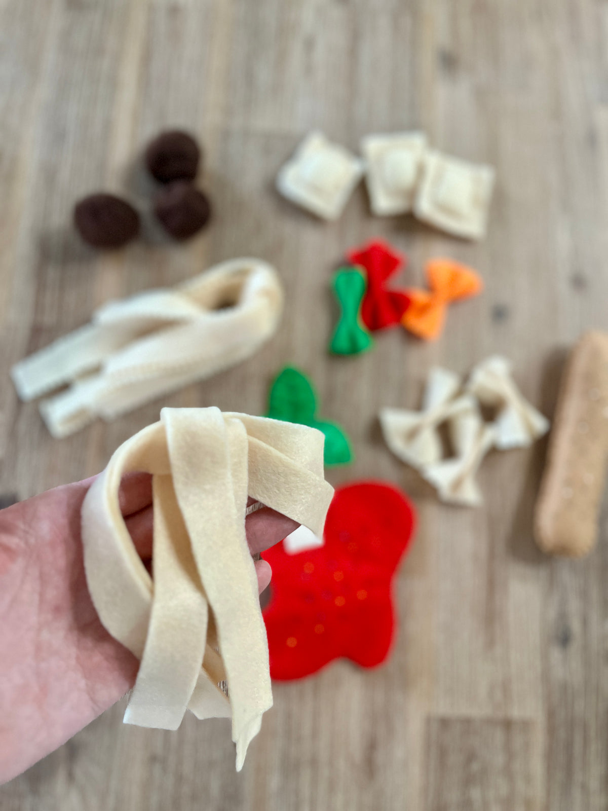 Close-up of a hand holding cream-colored felt pasta noodles with blurred felt pasta shapes including ravioli, farfalle, tomato slice, and sauce in the background, Felt Pasta Set, Made in Canada, handcrafted, homeschool, education resources.