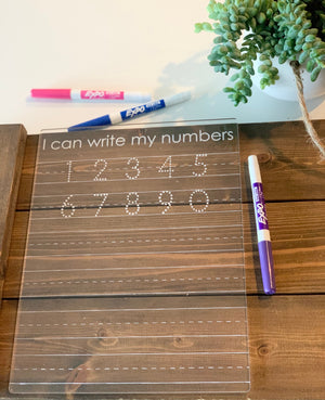 Transparent acrylic number tracing board with dotted digits and three colored dry erase markers on wooden table next to plant