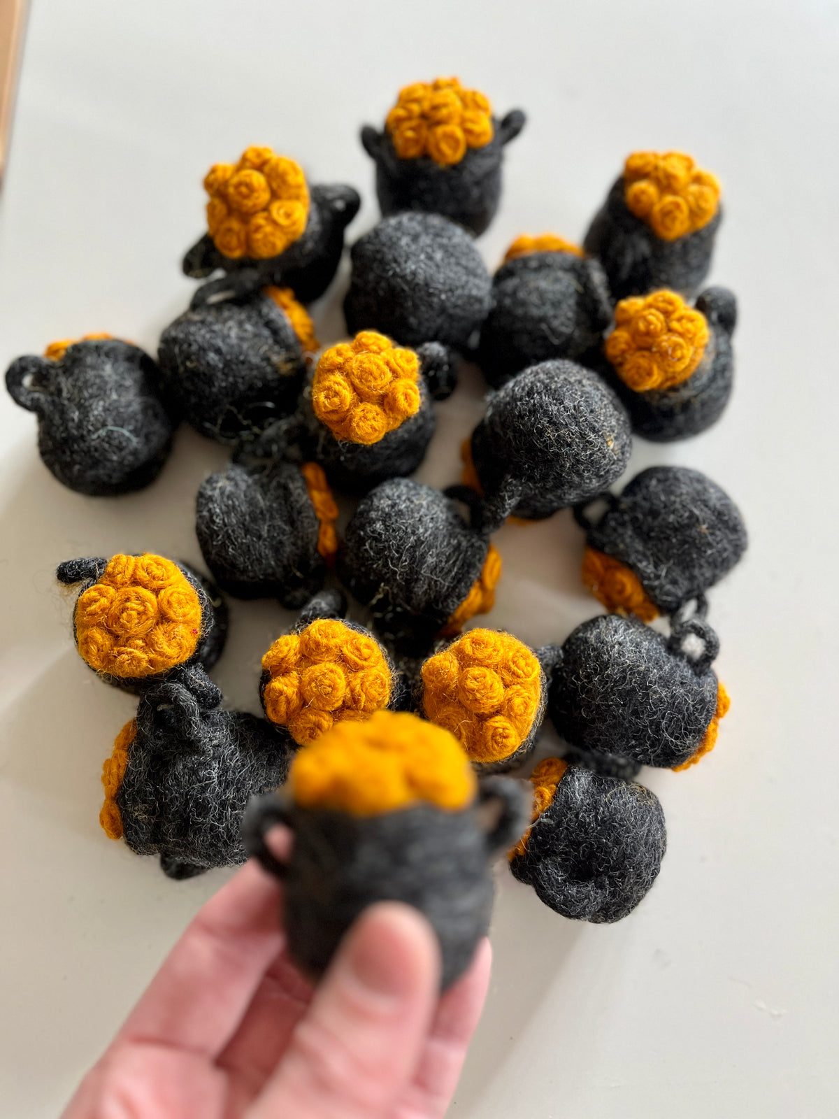 Hand holding small black felt pots topped with bright yellow rosette clusters scattered on white surface