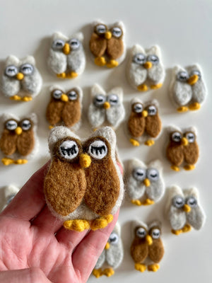 Hand holding a small brown and white felt owl figure with closed eyes above scattered similar felt owls on white background