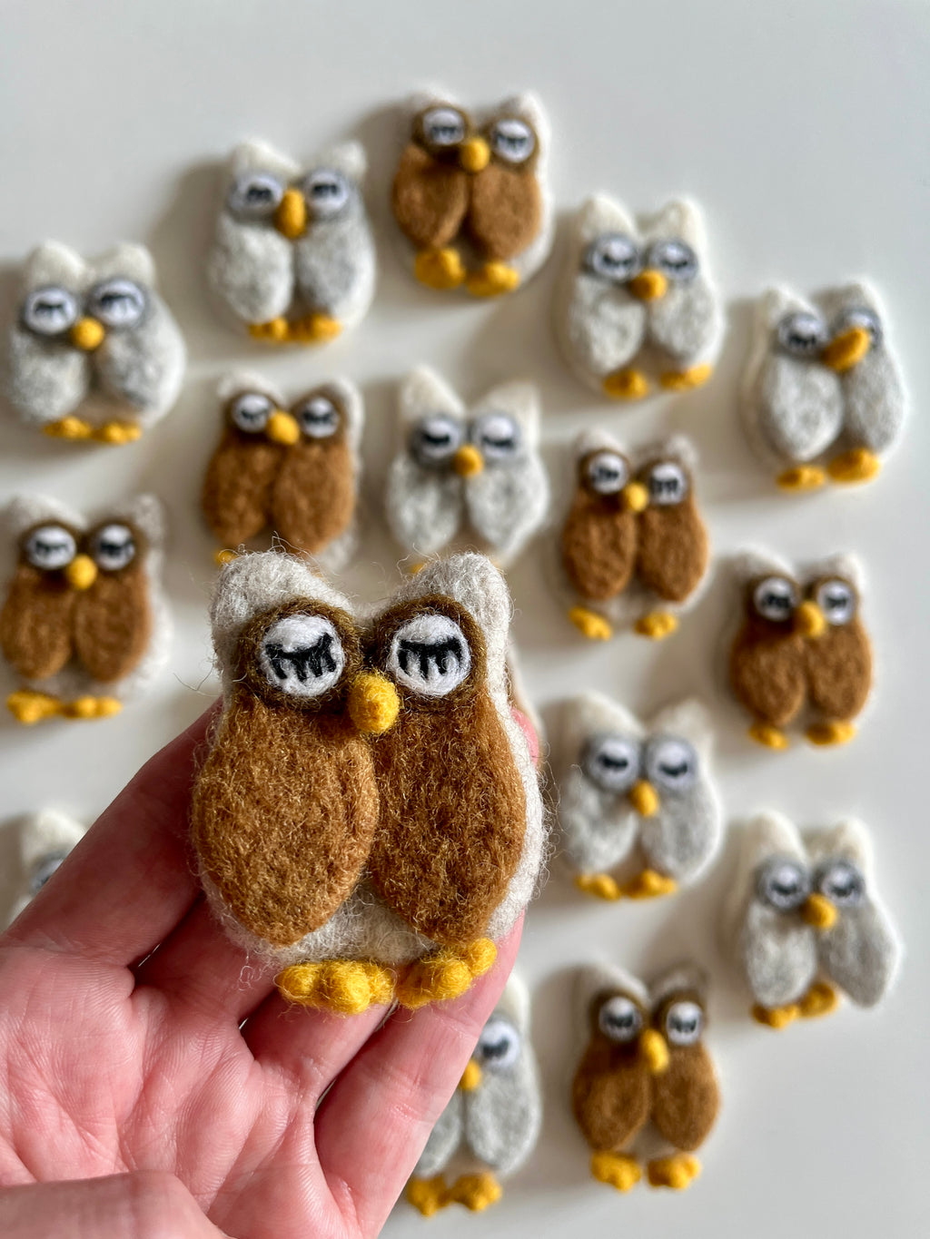 Hand holding a small brown and white felt owl figure with closed eyes above scattered similar felt owls on white background