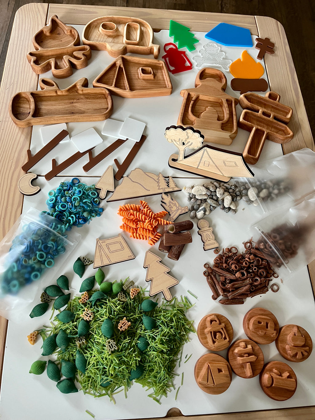 Wooden camping-themed trays and cutouts with trees, signs, campfire, tent, camper, colorful pasta, beads, and small natural elements on table