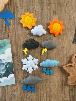 Colorful handcrafted felt weather shapes including suns, clouds with raindrops and lightning, and a snowflake, arranged on a wooden surface with children's books and trays, Felt Weather Shapes, Made in Canada, handcrafted, homeschool, education resources.